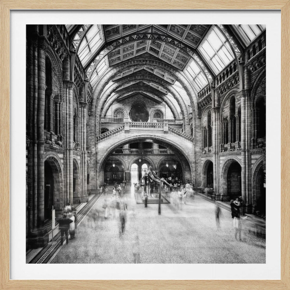 A black and white long exposure photograph of the grand hall of a natural history museum. The ornate, high-arched Romanesque architecture is in sharp focus, while a dinosaur skeleton stands in the center. Crowds of people are blurred into ghostly apparitions as they move through the vast space. Print