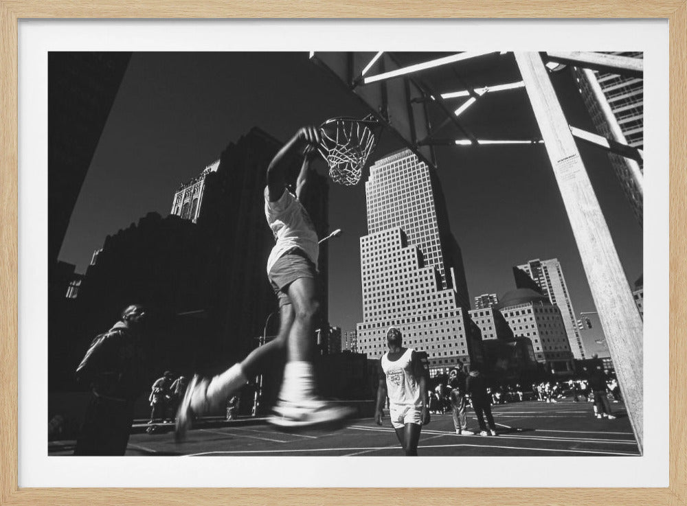 A low-angle, black and white action shot of a person dunking a basketball on an outdoor court, with tall city skyscrapers dominating the background under a clear sky. Print