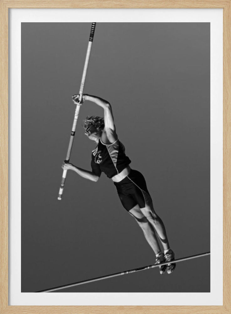 A dynamic black and white, low-angle shot of a female pole vaulter in mid-action. She is suspended in the air, gripping the pole as she prepares to clear the high bar, with her muscular form silhouetted against a plain sky. Print