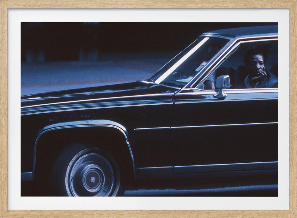 A moody, blue-toned photograph of a Black man sitting pensively in the driver's seat of a dark vintage sedan at night, framed in silver. Poster