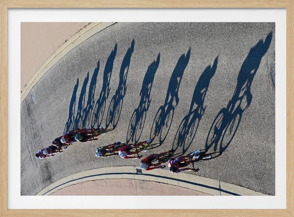 An aerial, high-angle photograph of a line of cyclists racing on a curved asphalt road. The bright sun casts long, dark, dramatic shadows of the riders and their bicycles, creating a striking diagonal pattern across the pavement. The image is framed in silver. Print