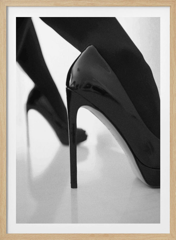 A close-up, black and white photograph of a person wearing shiny black patent leather stilettos and dark stockings. One shoe is in sharp focus in the foreground, highlighting its high arch and thin heel, while the other leg and shoe are softly blurred in the background on a reflective white surface. Decor