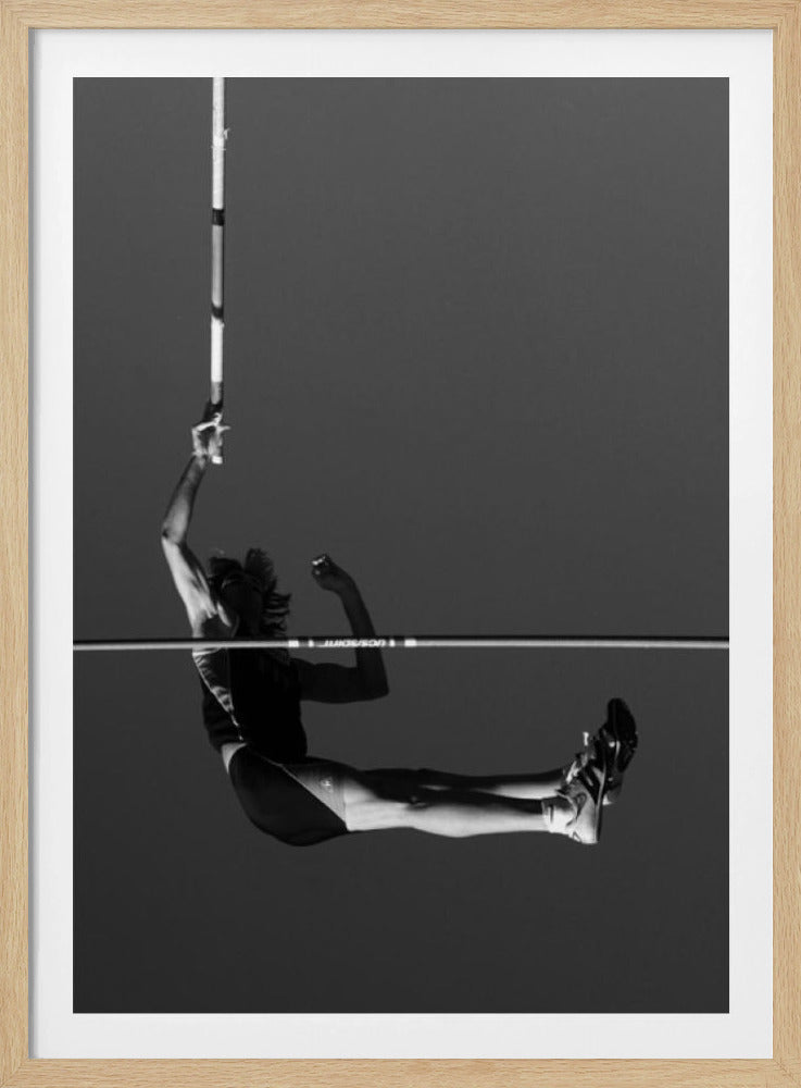 A dramatic black and white photograph of a male pole vaulter in mid-air, successfully clearing the horizontal bar. The athlete's body is parallel to the ground, with one arm still holding the vertical pole, against a solid gray sky. The image is enclosed in a simple black frame. Artwork