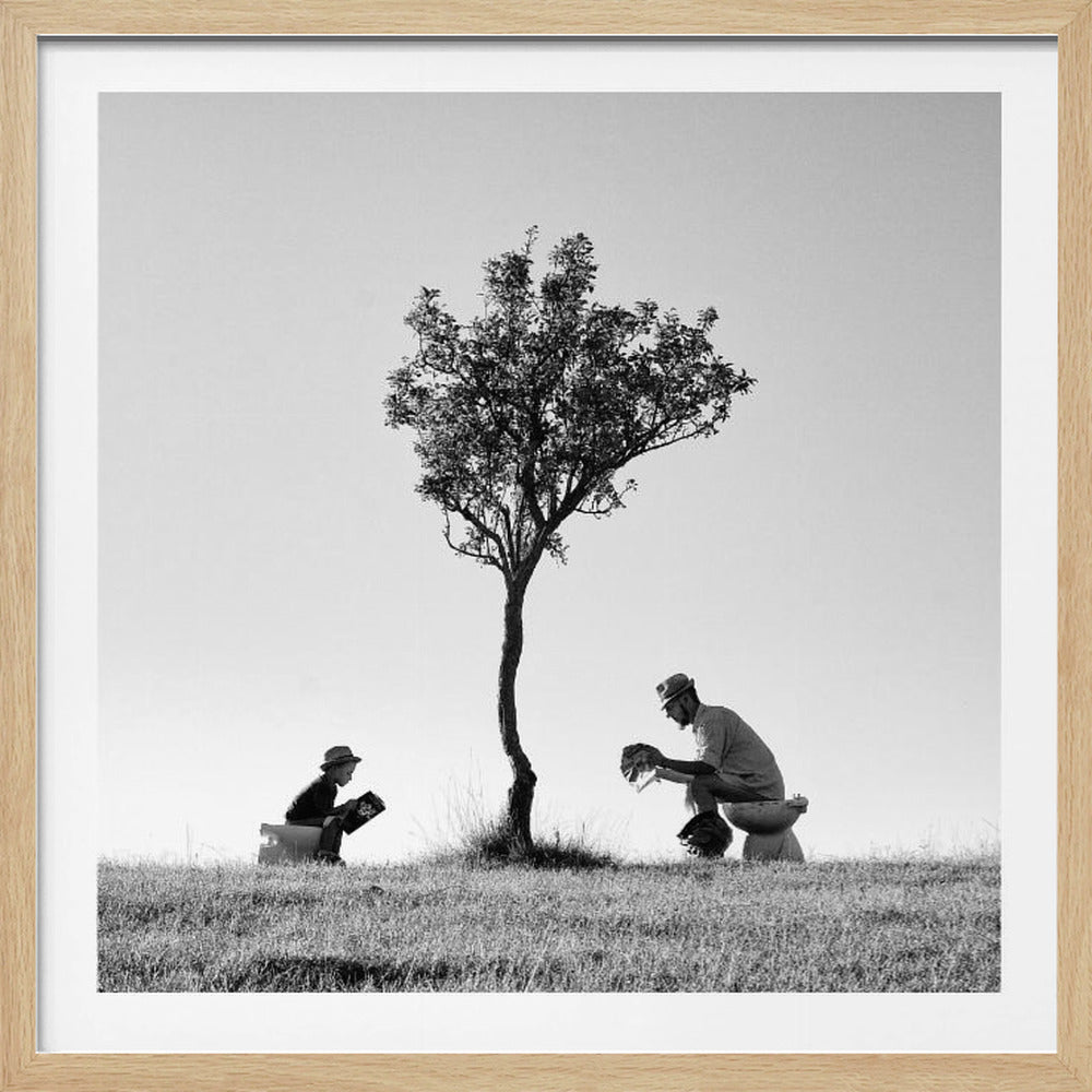 A framed, black and white photograph depicting a surreal scene of a man and a child sitting on toilets in an open grassy field under a single tree, both wearing hats and reading. Print