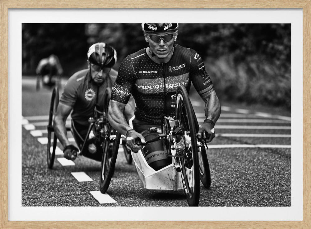 A powerful black and white photograph of two male athletes competing in a wheelchair race. The focused athlete in the foreground shows intense determination on his face, his muscular arms propelling the specialized racing wheelchair forward on an asphalt road. A second racer is visible in the background. Decor