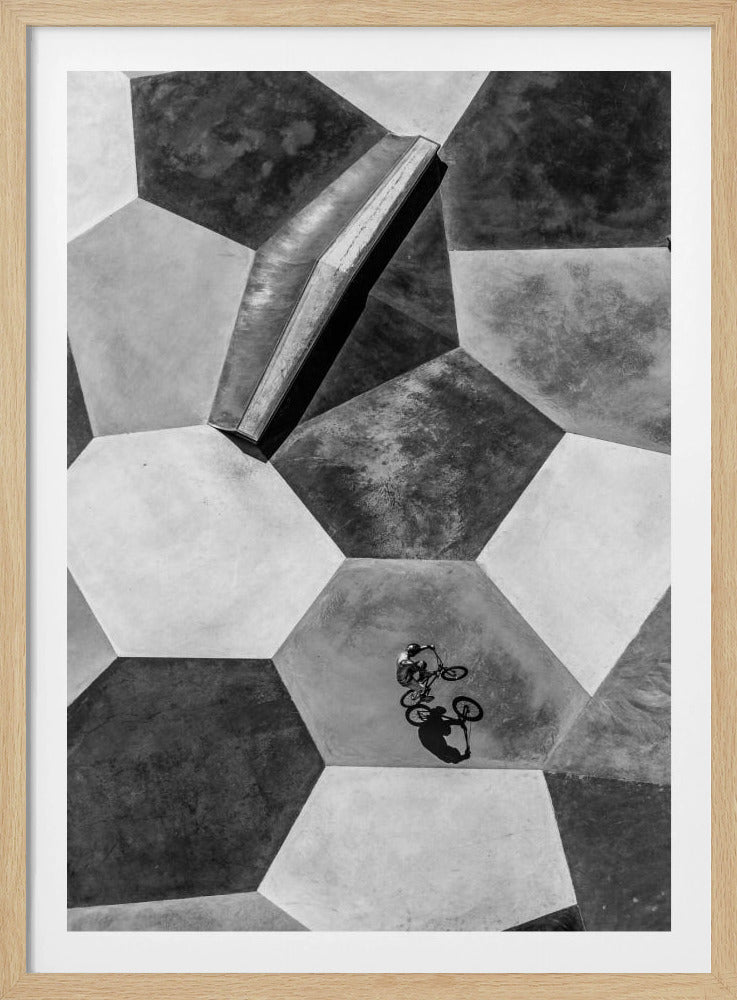 A high-angle, black and white photograph of a BMX rider in a skatepark with a geometric pattern. The ground consists of large, interlocking hexagons in various shades of grey, and the rider is captured mid-action on one of them, with a sharp shadow cast beside them. Artwork