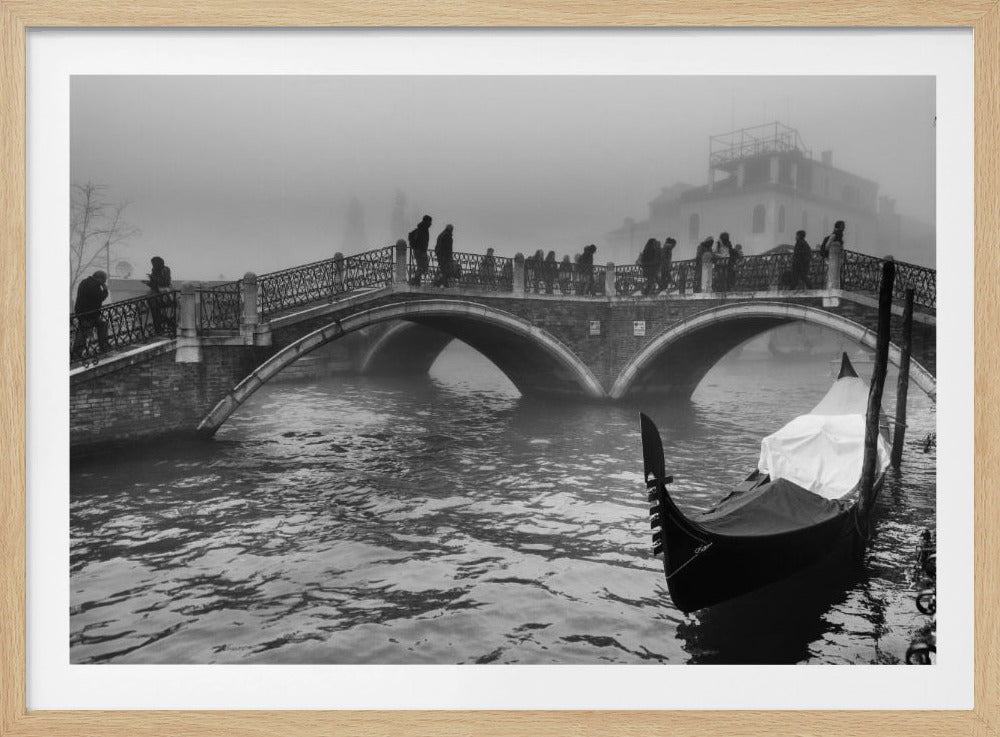A moody, black and white photograph of Venice on a foggy day. In the foreground, a gondola is moored in a rippling canal. In the background, silhouettes of people cross an arched stone bridge, and distant buildings are veiled in mist. The photo is displayed in a silver frame. Print
