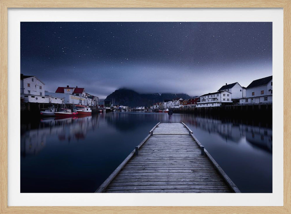 A long exposure photograph of a serene coastal town at night, viewed from a wooden pier that leads into calm, dark water. A lone person stands at the end of the pier, gazing at the town's reflected lights. Behind the village, a mountain looms under a breathtaking, star-filled sky. The image is presented within a silver-colored frame. Artwork