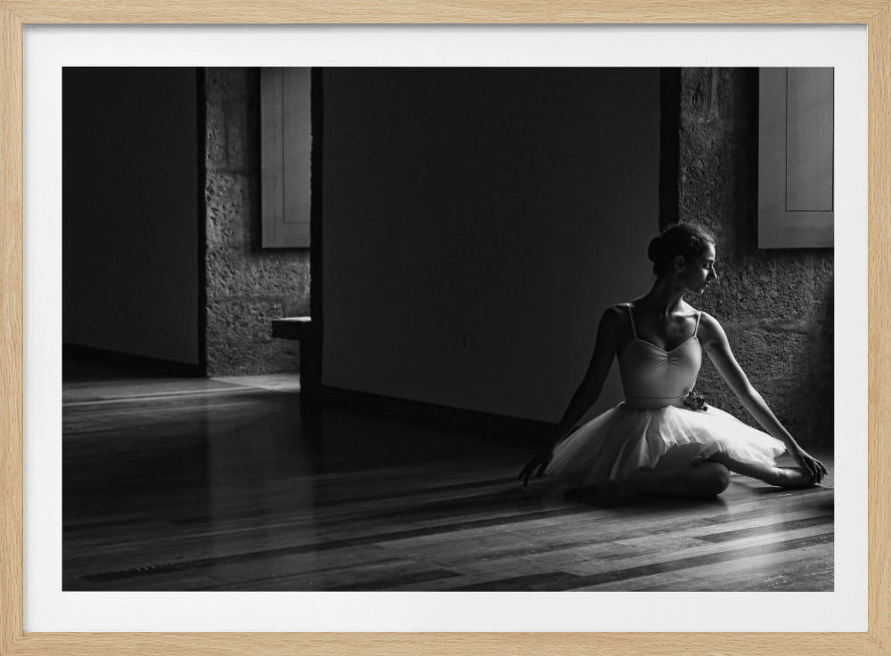 A dramatic black and white photograph of a ballerina sitting on a polished wooden floor in a dimly lit studio. She is wearing a white leotard and tutu, and a strong light from the side illuminates her, creating deep shadows and a contemplative mood. Wall Art
