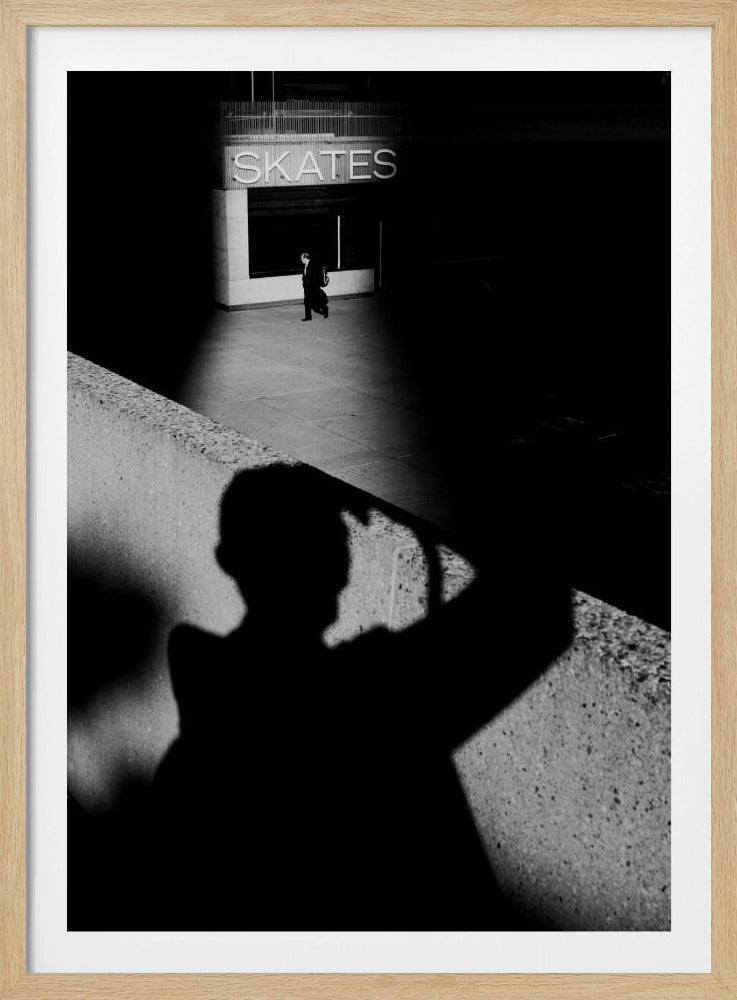 A high-contrast, black and white street photograph where the large shadow of the photographer is cast on a concrete barrier in the foreground. In the background, a lone person walks through a spotlit area of sidewalk in front of a building with an illuminated sign that says 'SKATES'. Poster