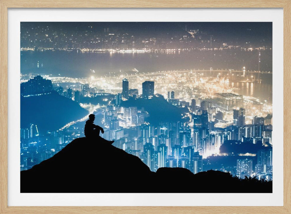 A silhouette of a person sitting on a hilltop, gazing out over a vast, illuminated city skyline at night. The city lights glow in shades of blue and gold against the dark landscape, with a body of water in the distance. Decor