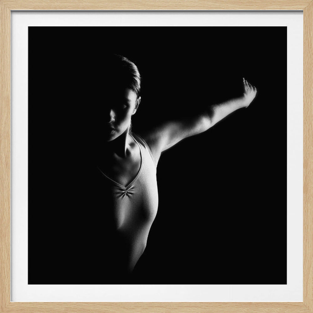 A dramatic black and white studio photograph of a dancer in a leotard against a black background. Strong side lighting illuminates her form and extended arm, while her face remains mostly in shadow, creating a high-contrast, artistic effect. The image is presented in a light wood frame. Decor
