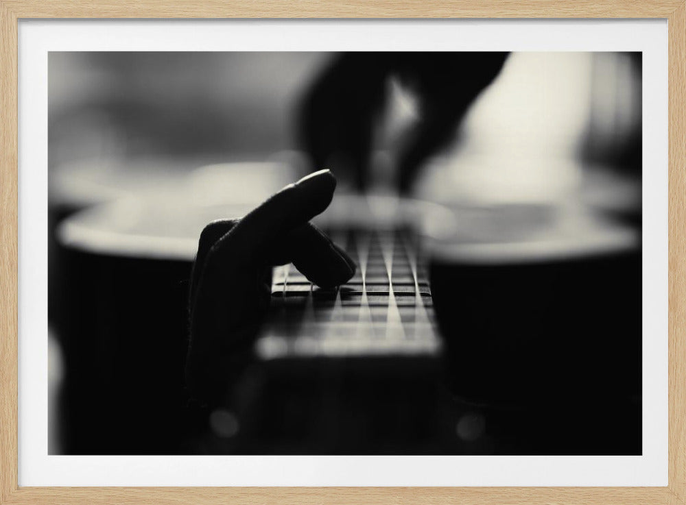 A dramatic black and white close-up shot of a person's hand pressing strings on the fretboard of a guitar. The focus is sharp on the fingers and frets, with the rest of the guitar and the strumming hand blurred in the background, all enclosed in a silver frame. Decor