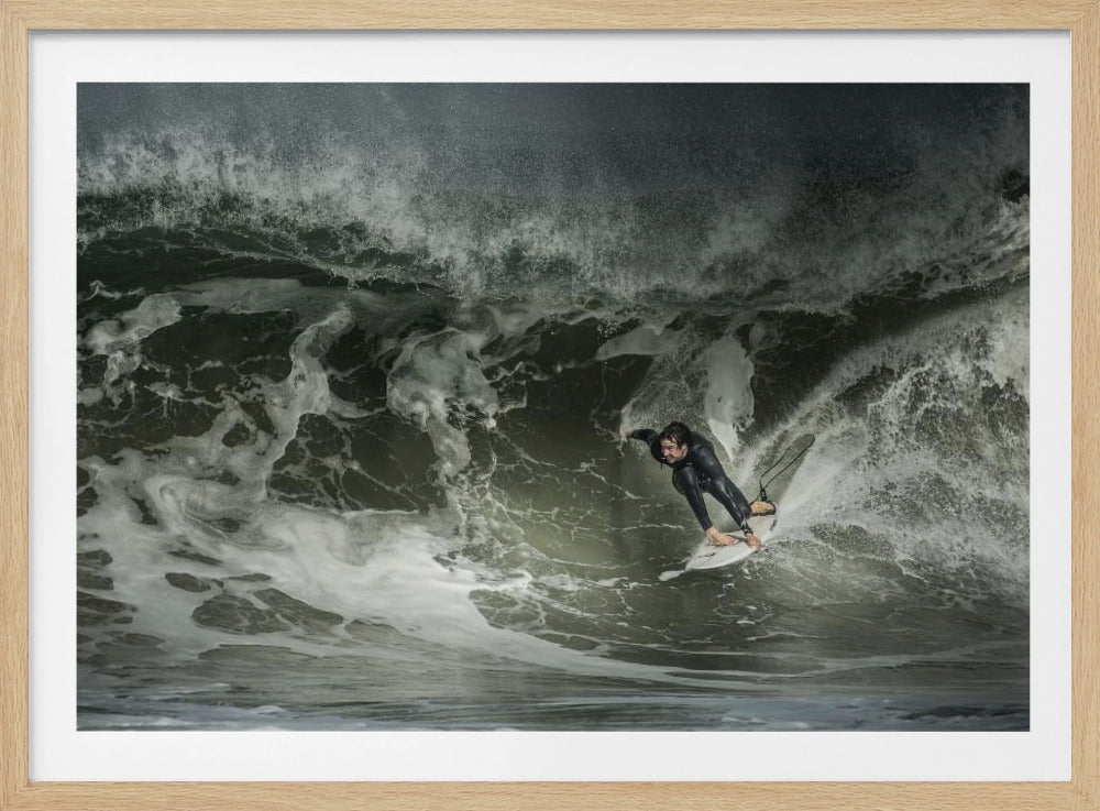 A dramatic overhead photograph of a surfer in a black wetsuit riding a white surfboard on a large, dark, foamy wave. The scene is captured in a moody, almost monochromatic tone and is presented within a silver picture frame. Artwork