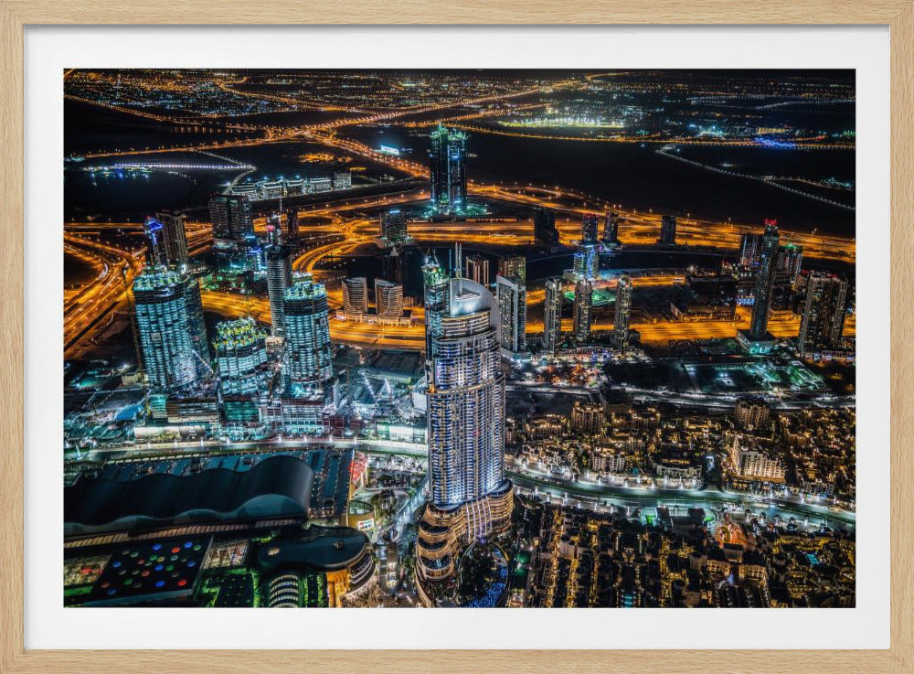 An aerial nighttime photograph of a sprawling modern city, possibly Dubai, with brightly lit skyscrapers and a complex network of illuminated highways creating a vibrant contrast against the dark landscape, all enclosed in a silver frame. Decor