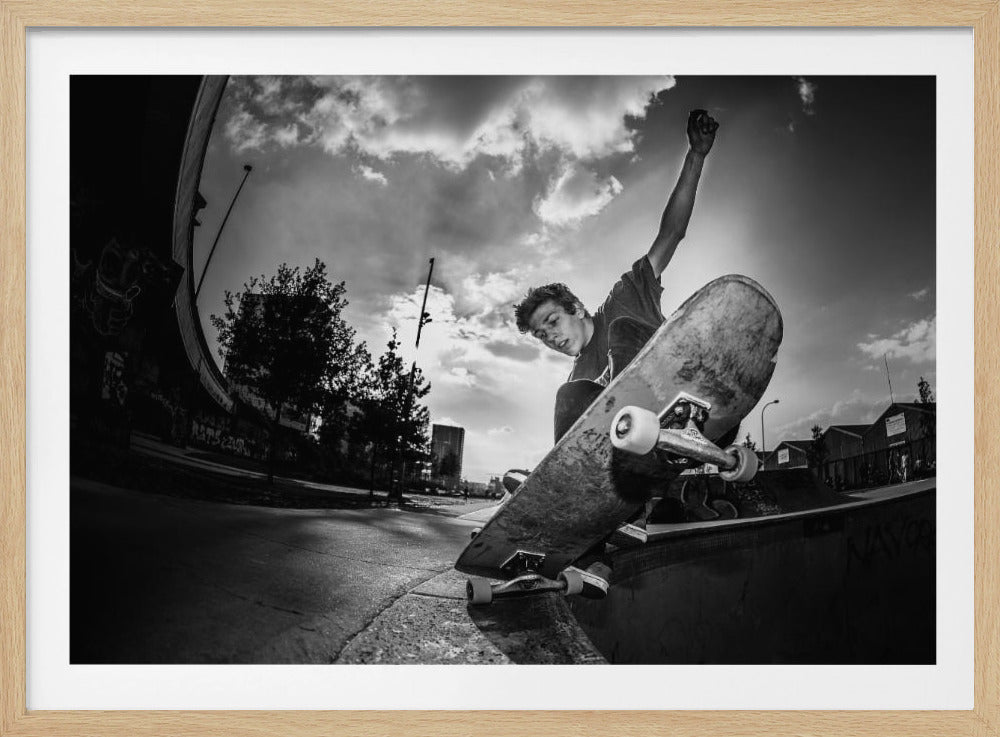 A dynamic, low-angle, black and white photograph of a young man skateboarding. He is balancing on the edge of a ramp with his skateboard angled up towards the viewer. He has one arm raised in the air with a clenched fist. The shot is taken with a fisheye lens, curving the horizon against a dramatic cloudy sky in an urban setting. The image is presented within a silver frame. Decor