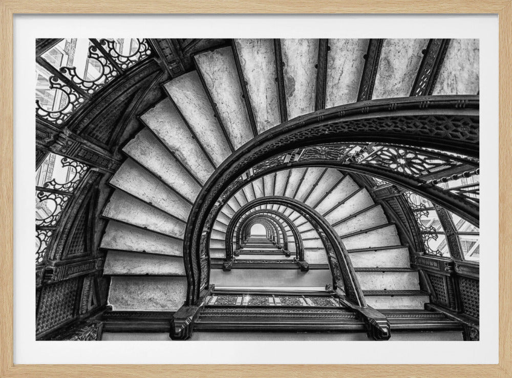 A dramatic black and white photograph looking up from the bottom of an ornate spiral staircase. The dark, intricate ironwork of the railings contrasts with the light stone steps, creating a mesmerizing, repeating pattern that spirals towards a bright light at the top. The image is presented within a silver-colored frame. Decor