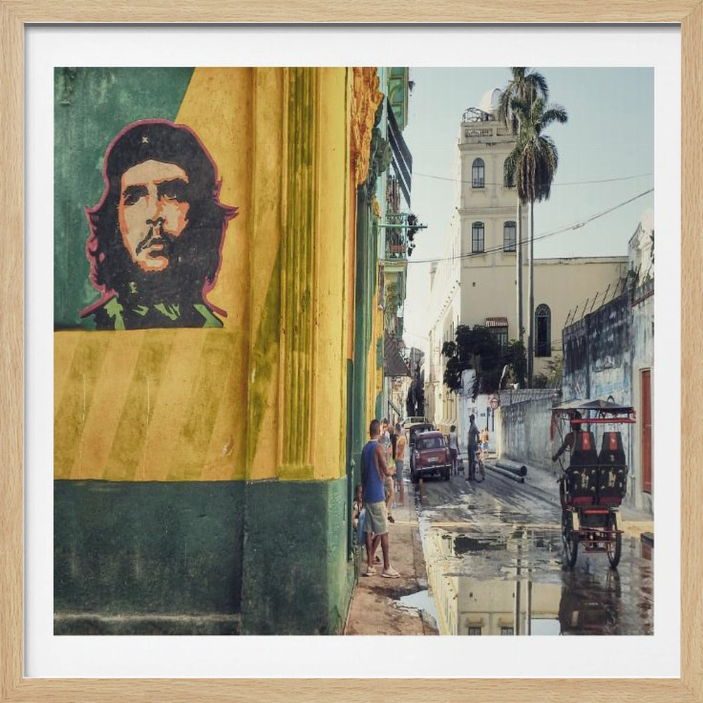 A framed photograph of a vibrant street corner in a city like Havana, Cuba, with a large mural of Che Guevara on a yellow and green wall. The wet street reflects the buildings and a palm tree, while people, a vintage car, and a rickshaw occupy the narrow road. Poster