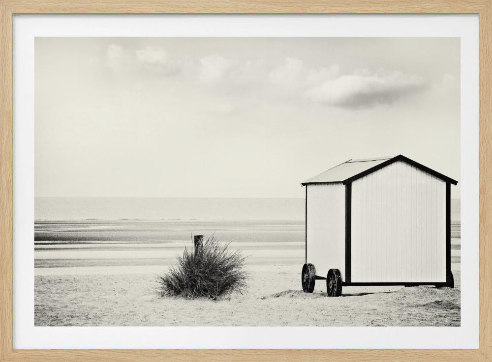 A black and white photograph of a tranquil beach scene. On the right, a small, white beach hut with black trim and wheels sits on the sand. In the background, the calm sea stretches to the horizon under a pale, cloudy sky. Wall Art