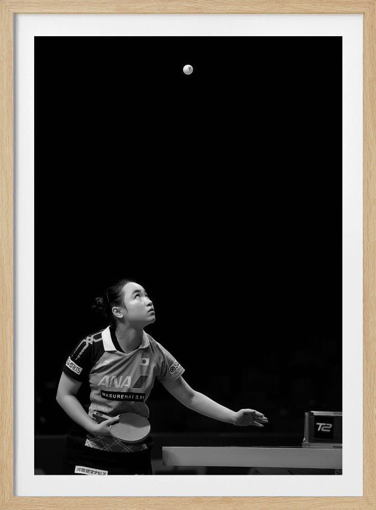 A dramatic black and white vertical photograph of a female table tennis player mid-serve. Positioned in the lower-left, she looks up with intense focus at the white ball suspended high in the air against a solid black background. Poster