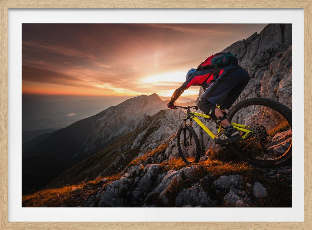 A mountain biker in full gear rides a yellow bike down a narrow, rocky mountain ridge at sunset. The sky is filled with the warm colors of the setting sun, which illuminates the distant mountain peaks. Artwork