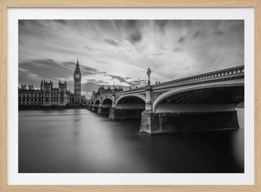 A dramatic black and white long-exposure photograph of London, featuring the smooth River Thames in the foreground, Westminster Bridge on the right, and the iconic Big Ben and Houses of Parliament on the left under a cloudy, streaked sky. The image is presented in a silver frame. Print