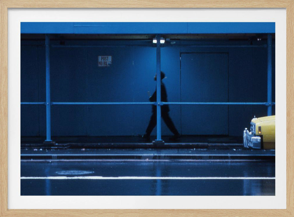 A moody, grainy photograph dominated by shades of blue, capturing the blurred silhouette of a person walking under a covered city sidewalk at night. The wet street in the foreground reflects light, and the yellow cab of a truck is partially visible on the right. Artwork