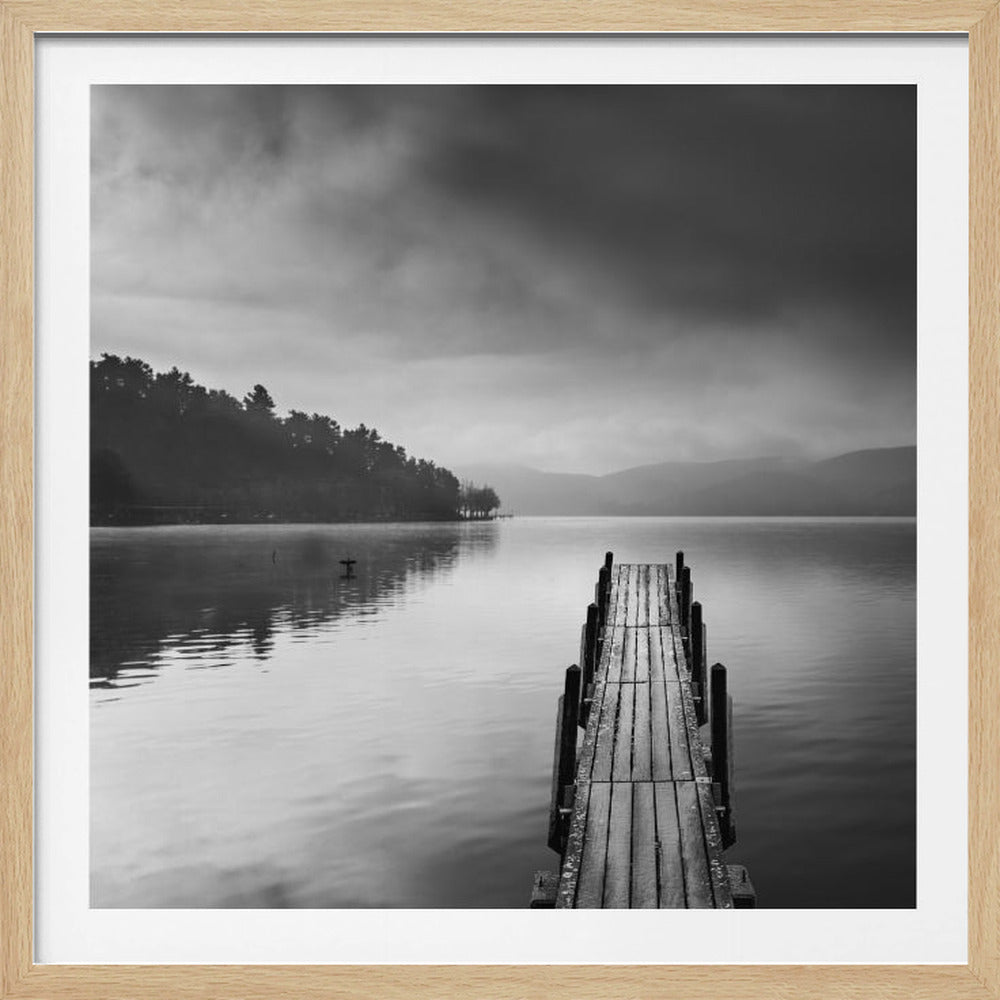 A framed, moody black and white photograph of a wooden pier extending into a calm lake under a cloudy sky, with a forested shoreline and distant mountains in the background. Poster