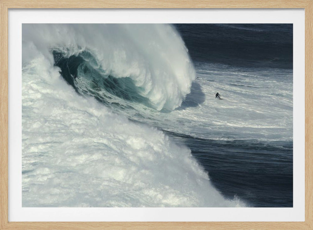 A framed aerial photograph captures a dramatic ocean scene where a lone surfer on a surfboard is dwarfed by an immense, powerful wave cresting nearby, conveying a sense of scale and adventure. Artwork