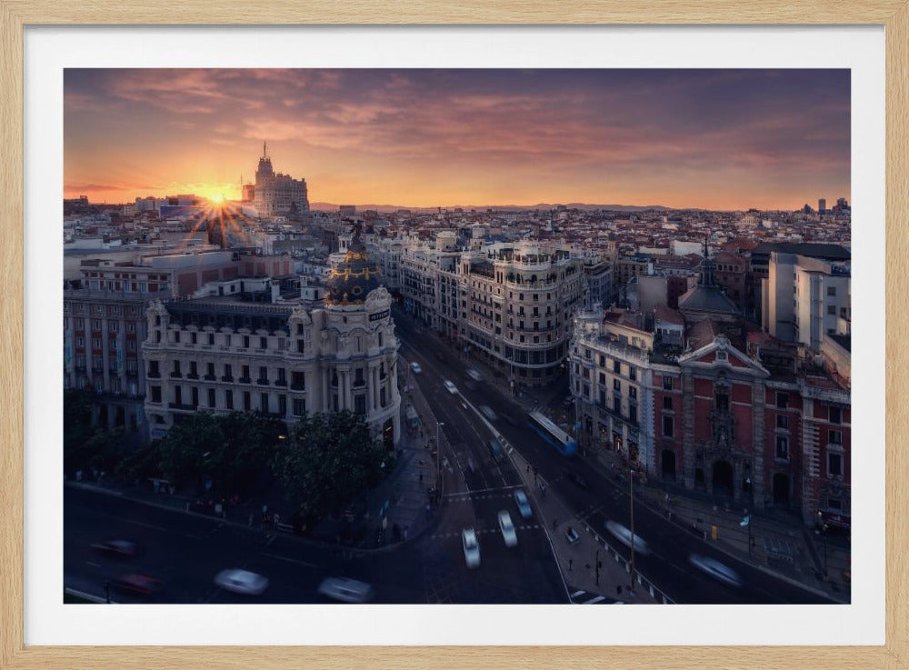A framed aerial photograph of the Madrid skyline at sunset, featuring the iconic Metropolis Building and blurred traffic on the streets below, under a vibrant orange and purple sky. Decor