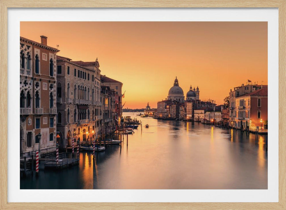 A scenic photograph of the Grand Canal in Venice at sunset, viewed from a high vantage point and enclosed in a silver frame. The sky is a vibrant orange, casting a warm, golden light on the historic buildings lining the waterway and reflecting on the smooth surface of the water. In the distance, the iconic domes of the Basilica di Santa Maria della Salute are silhouetted against the glowing sky. Artwork