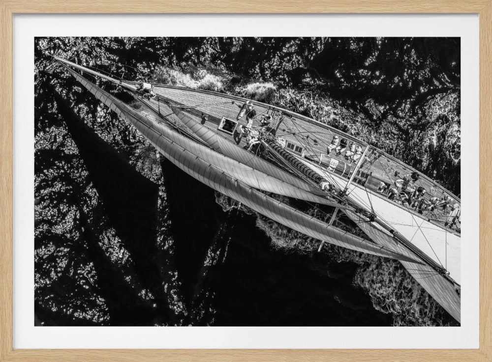 A dramatic black and white overhead aerial photograph of a large sailboat cutting through dark, choppy water. The crew is visible on the wooden deck, managing the full sails as the yacht heels over, creating a white wake. The image is presented within a silver frame. Print