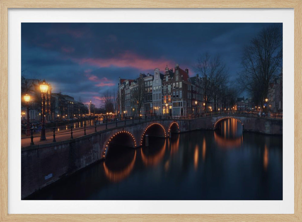 A tranquil evening cityscape of a canal in Amsterdam, with a multi-arched brick bridge illuminated by golden lights that reflect beautifully on the dark, still water. The sky above is a deep twilight blue with streaks of pink from the sunset, and historic buildings line the waterway. Wall Art