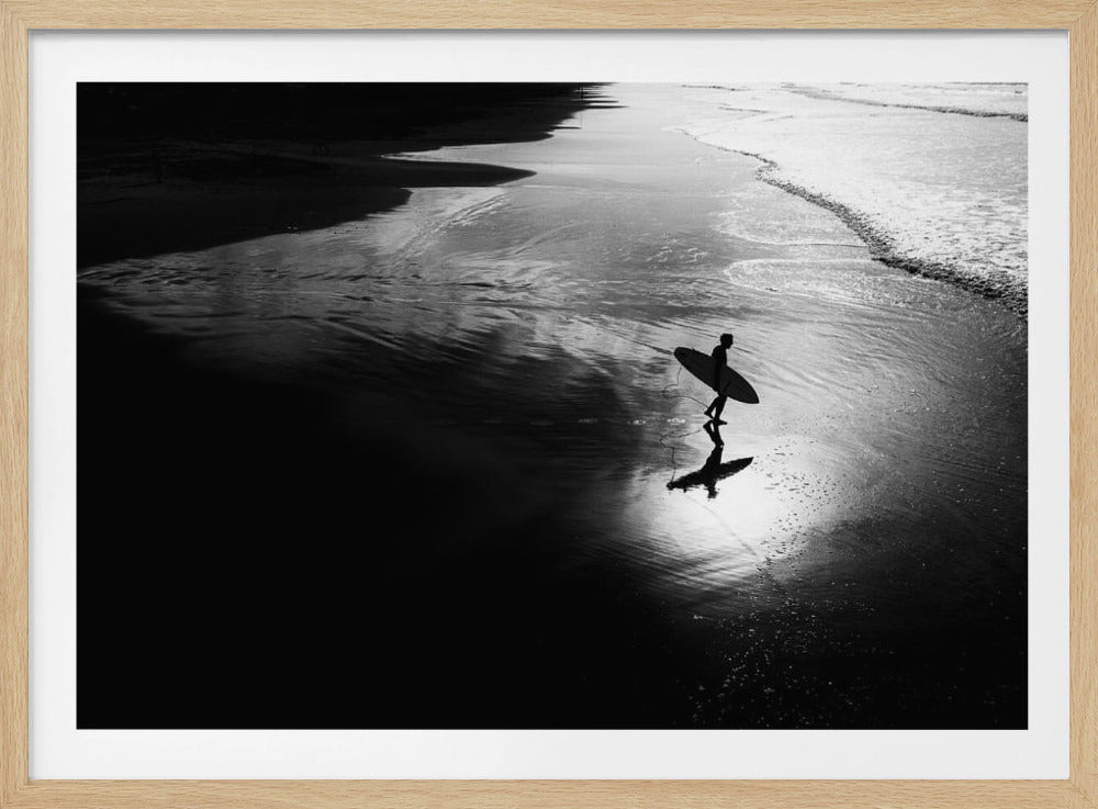 A high-contrast, black and white photograph of a silhouetted surfer carrying a surfboard and walking along the wet, reflective sand at the water's edge. Decor