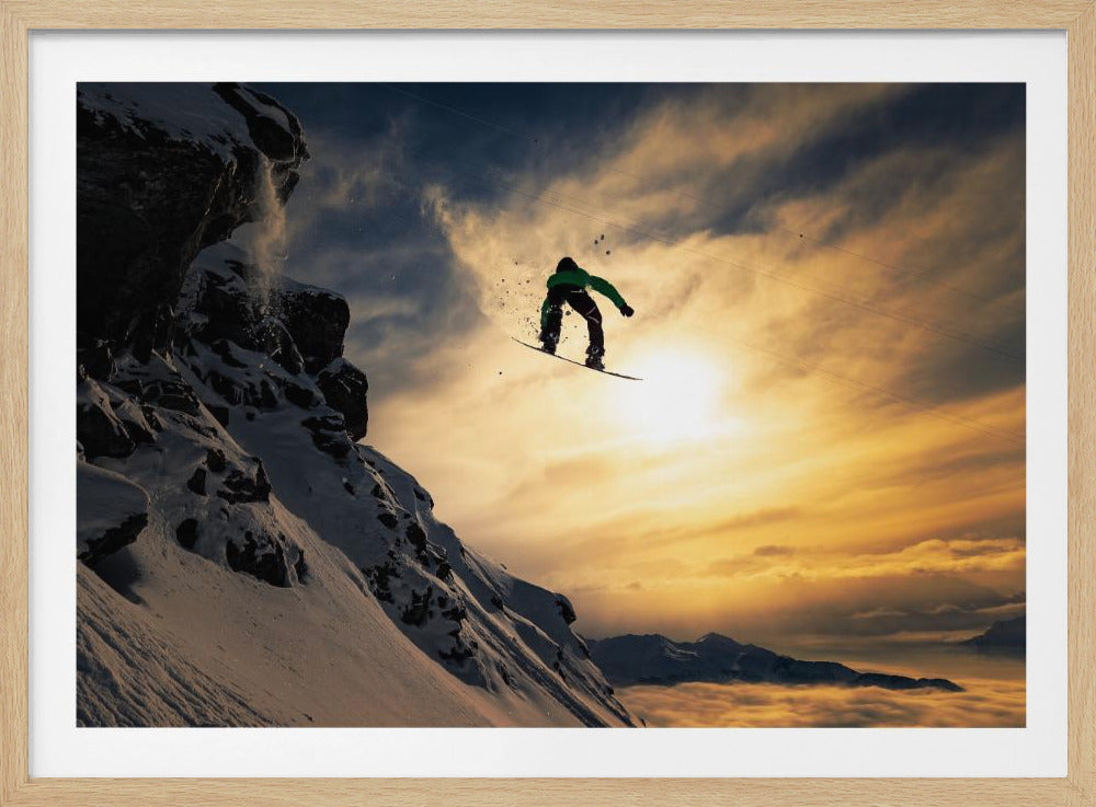 A snowboarder is captured in mid-air, silhouetted against a brilliant sunset after jumping off a snowy, rocky cliff. Below the action, a stunning mountain range peeks through a thick layer of clouds, illuminated by the golden hour light. Print