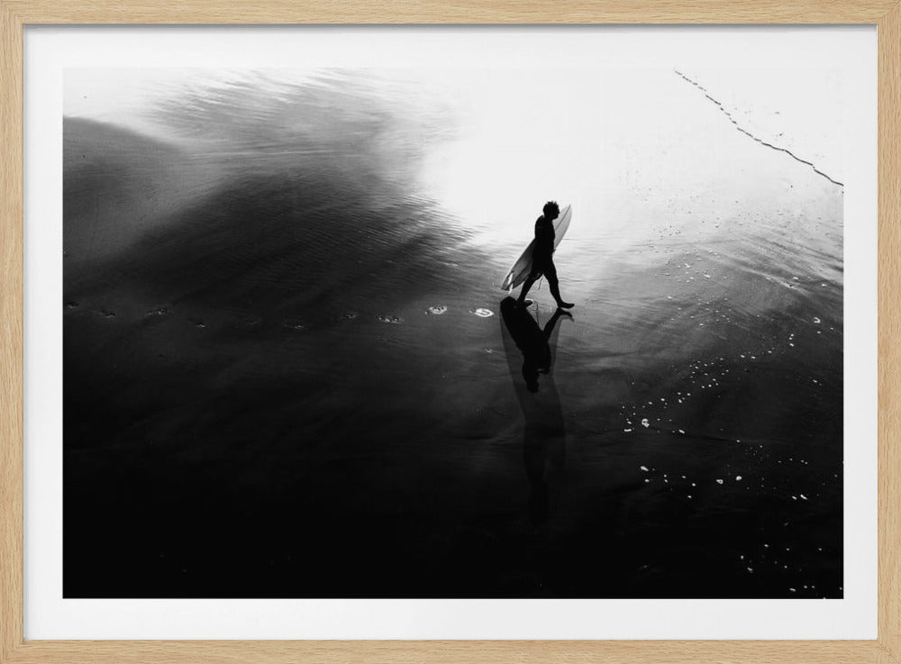 A high-contrast, black and white photograph of a surfer silhouetted against the bright reflection of the sun on the water. The surfer is carrying a surfboard and walking along the wet sand, leaving footprints behind. Their long shadow is cast in front of them on the dark, glistening sand. Print