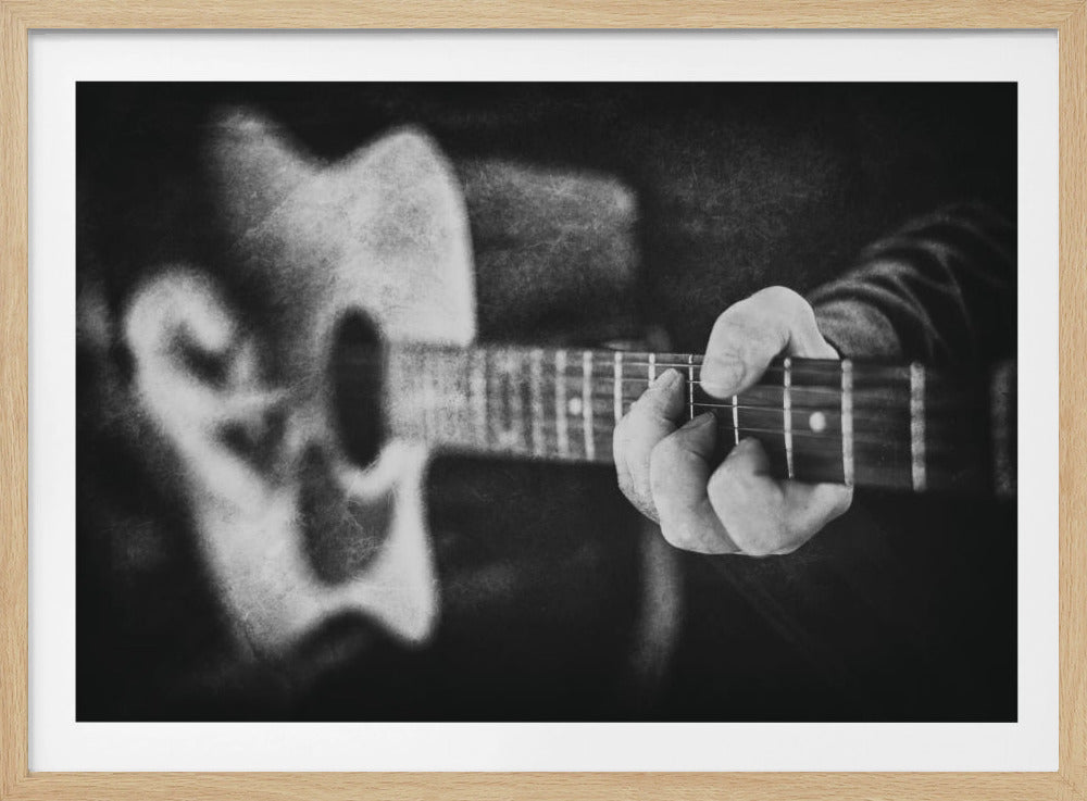 A grainy, black and white close-up photograph focusing on a person's hand fretting a chord on a guitar neck. The body of the guitar is softly blurred in the background, creating an artistic and moody feel. The image is presented within a silver frame. Print
