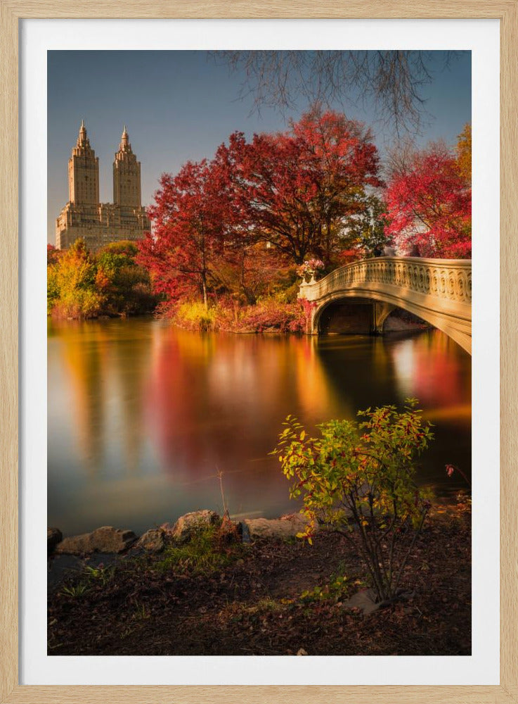 A picturesque autumn scene in Central Park featuring the Bow Bridge over a calm lake reflecting the vibrant red and yellow fall foliage, with the twin towers of The San Remo building visible in the background against a clear blue sky. Decor