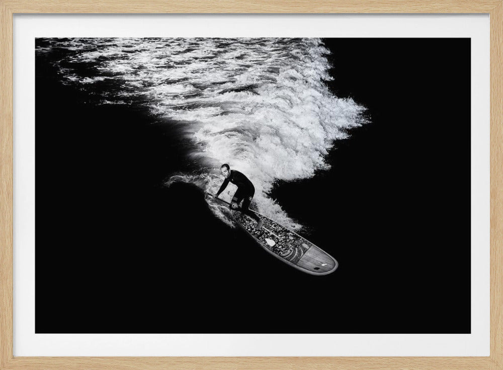 A dramatic black and white high-angle photograph of a man in a wetsuit paddleboarding on a wave, creating a stark white wake against the deep black water, all enclosed in a silver frame. Print