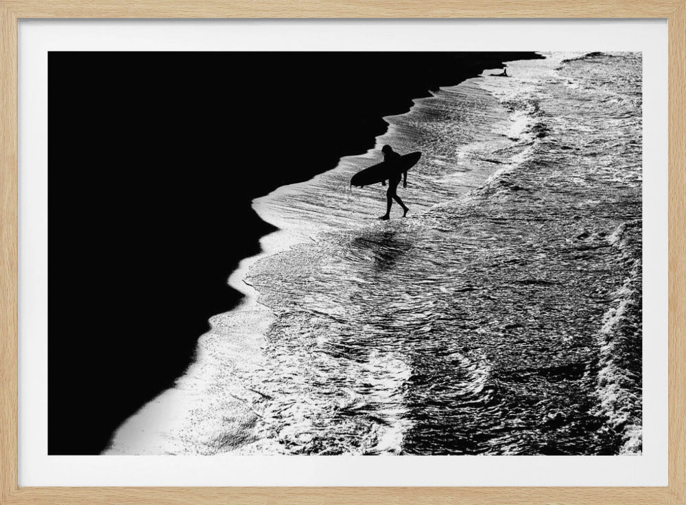 A high-contrast, black and white aerial photograph of a silhouetted surfer carrying a surfboard and walking along the shoreline. The image is divided diagonally, with the dark beach on the left and the white, foamy ocean waves on the right, creating a dramatic and graphic composition. The image is enclosed in a silver frame. Wall Art