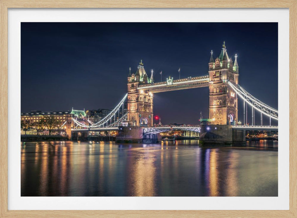 A stunning night photograph of London's iconic Tower Bridge, fully illuminated with golden and white lights that cast a shimmering reflection on the dark waters of the River Thames below, all set against a deep night sky and enclosed in a silver frame. Poster