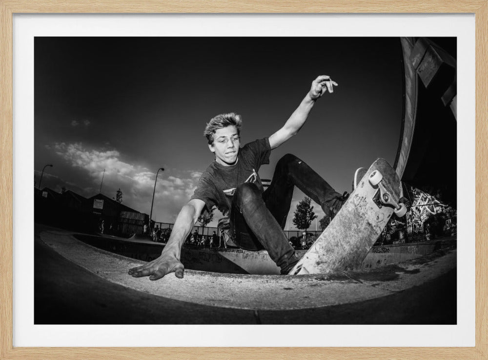 A dynamic, low-angle black and white action shot of a young skateboarder performing a trick in a concrete skatepark. He balances with one hand on the edge of the bowl, his body low to the ground as he carves along the curve, with his other arm raised in the air. Wall Art