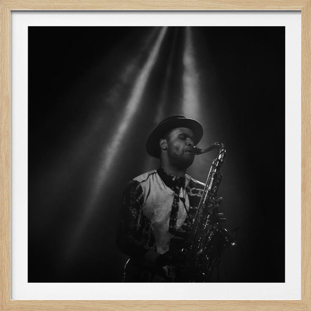 A dramatic black and white photograph of a musician wearing a fedora, playing the saxophone under spotlights. Beams of light cut through the dark background, highlighting the man as he plays with his eyes closed in concentration. The photo is displayed within a light-colored wooden frame. Decor