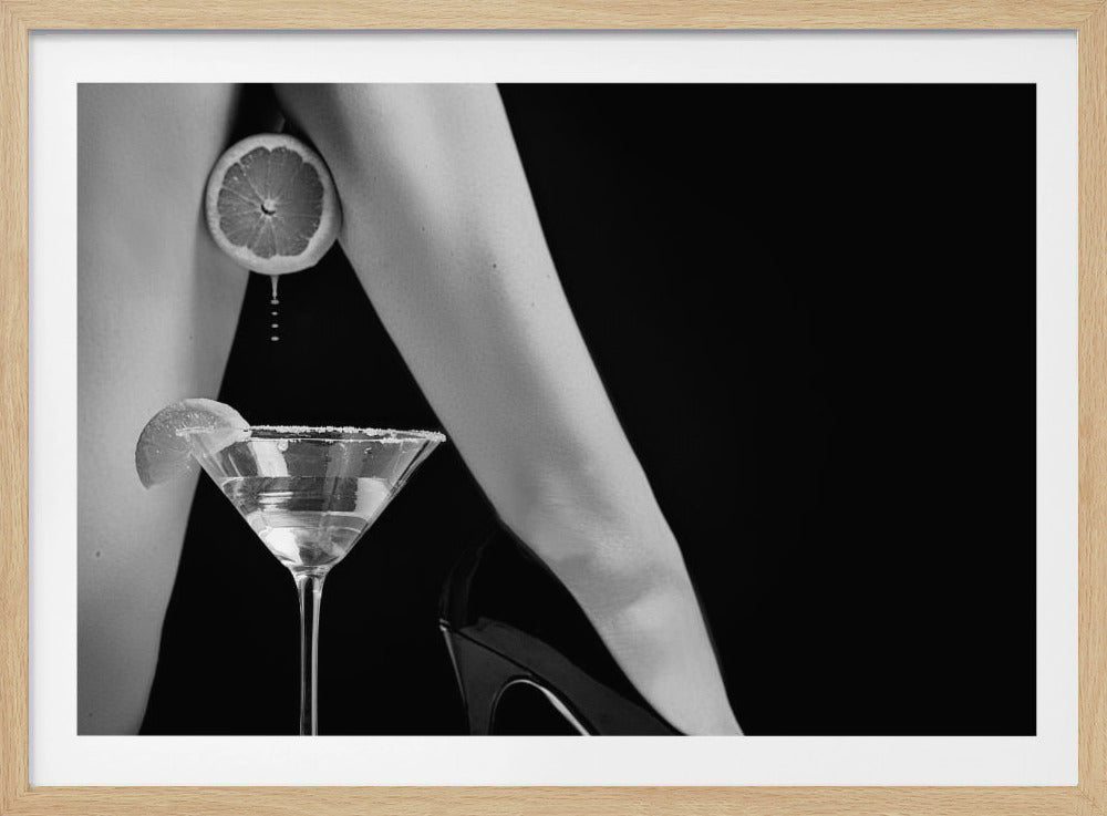 A framed, black and white photograph showing a woman's legs and a martini glass against a black background. A sliced citrus fruit is held between her thighs, with juice dripping towards the sugar-rimmed cocktail glass below. One foot is visible, wearing a black stiletto high heel. Artwork