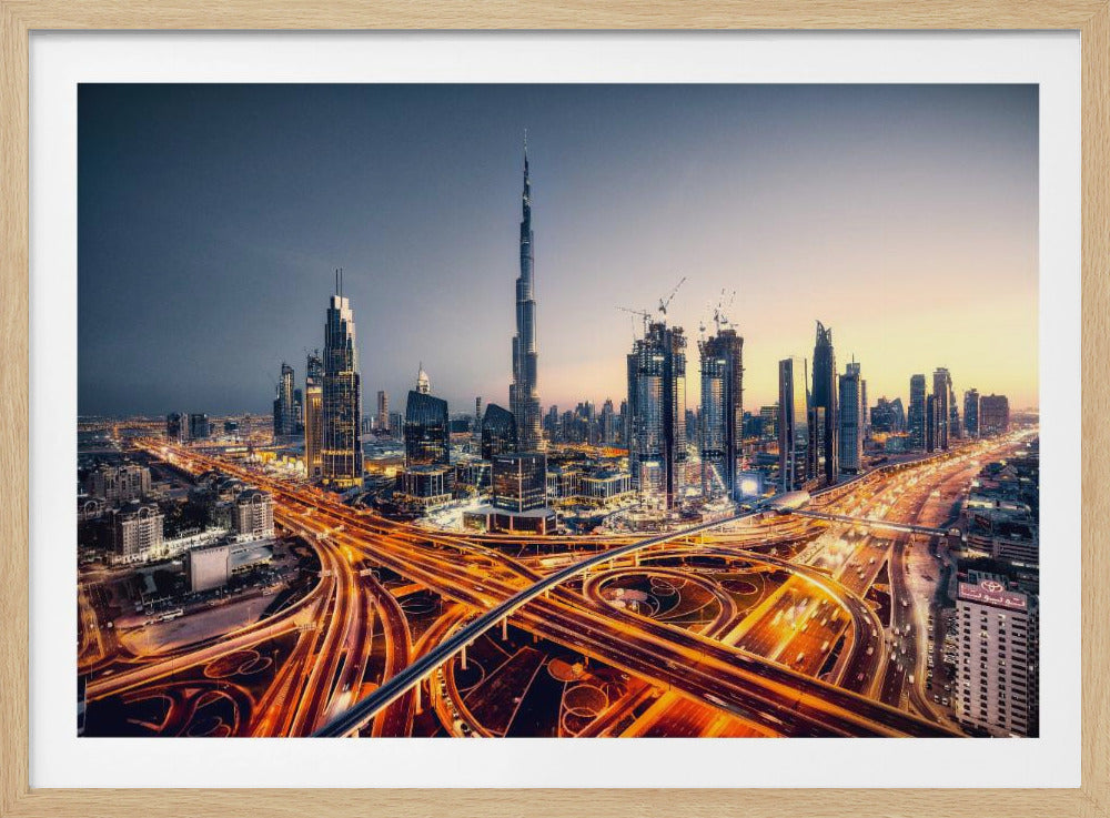 A wide-angle, elevated photograph of the Dubai skyline at dusk, with the iconic Burj Khalifa towering in the center. In the foreground, a complex network of highways glows with the orange light trails of moving traffic, while the city's numerous skyscrapers are illuminated against a twilight sky. Print