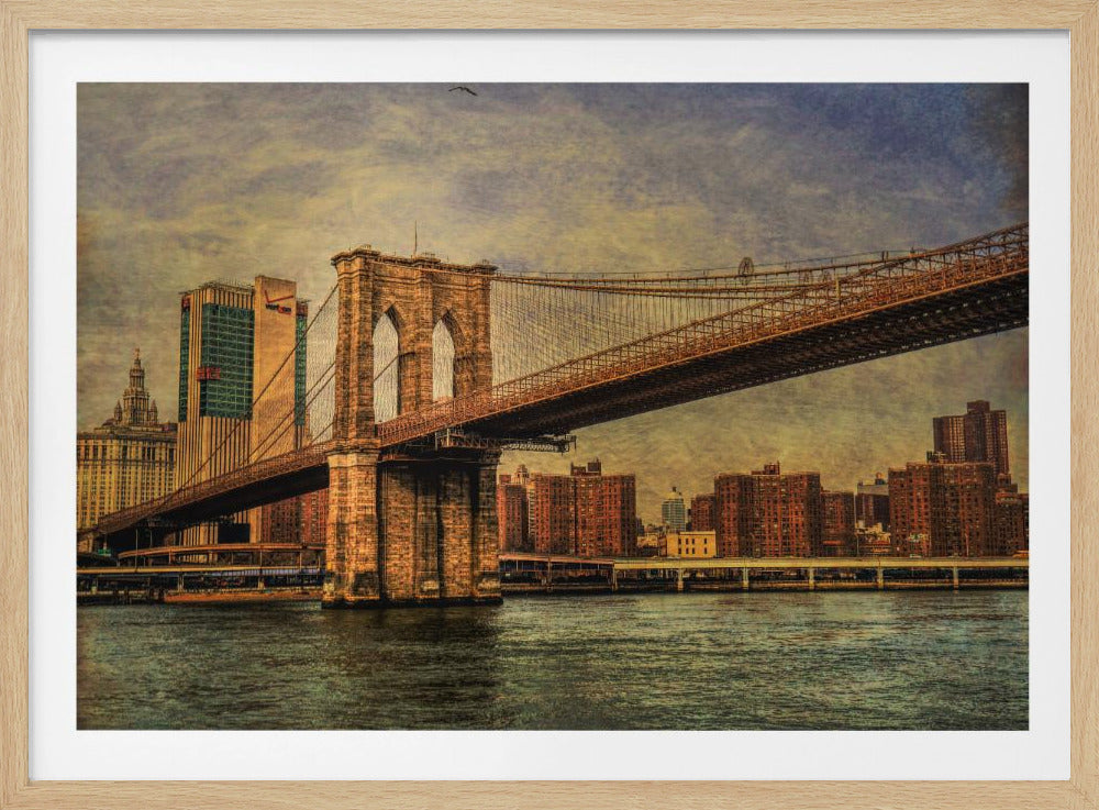 A framed, vintage-style photograph of the Brooklyn Bridge from a low angle, showing its stone pylon and suspension cables against a textured, cloudy sky. The Manhattan skyline and the East River are visible in the background and foreground. Wall Art