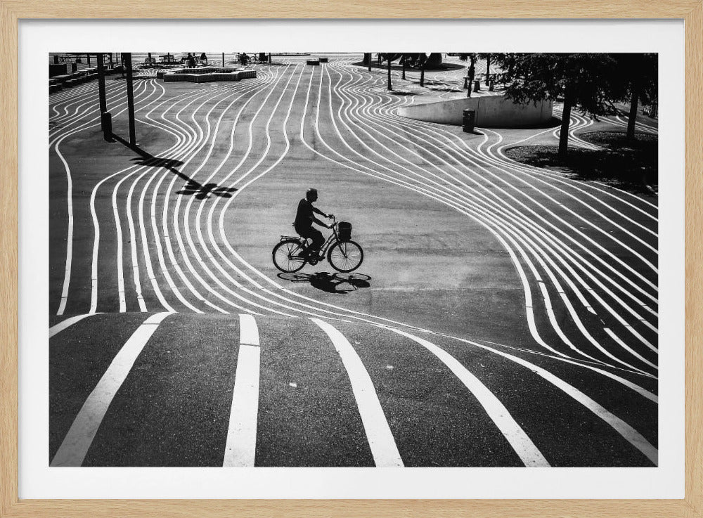 A high-contrast, black and white overhead photograph of a person riding a bicycle across a wide, paved plaza. The ground is covered in a mesmerizing pattern of wavy, parallel white lines, and the cyclist's long shadow is cast by the bright sun. Print