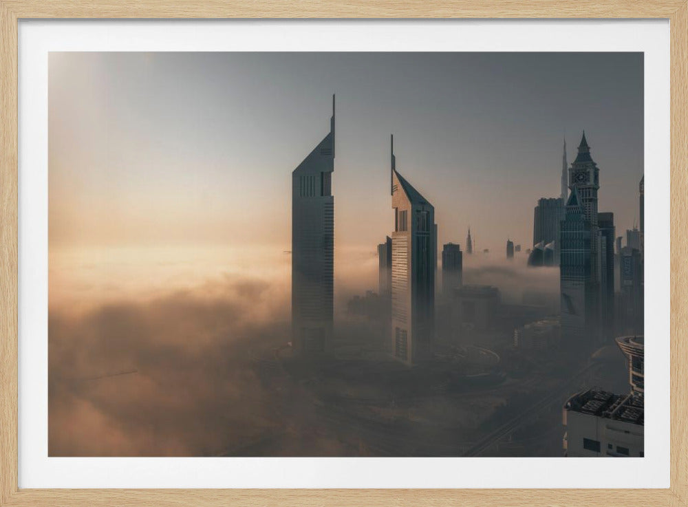 A framed aerial photograph of a futuristic city skyline, likely Dubai, enveloped in a thick layer of fog at sunrise. Tall, modern skyscrapers, including the iconic Emirates Towers, pierce through the clouds, with the warm light of the rising sun creating a hazy, golden glow on the left. Poster