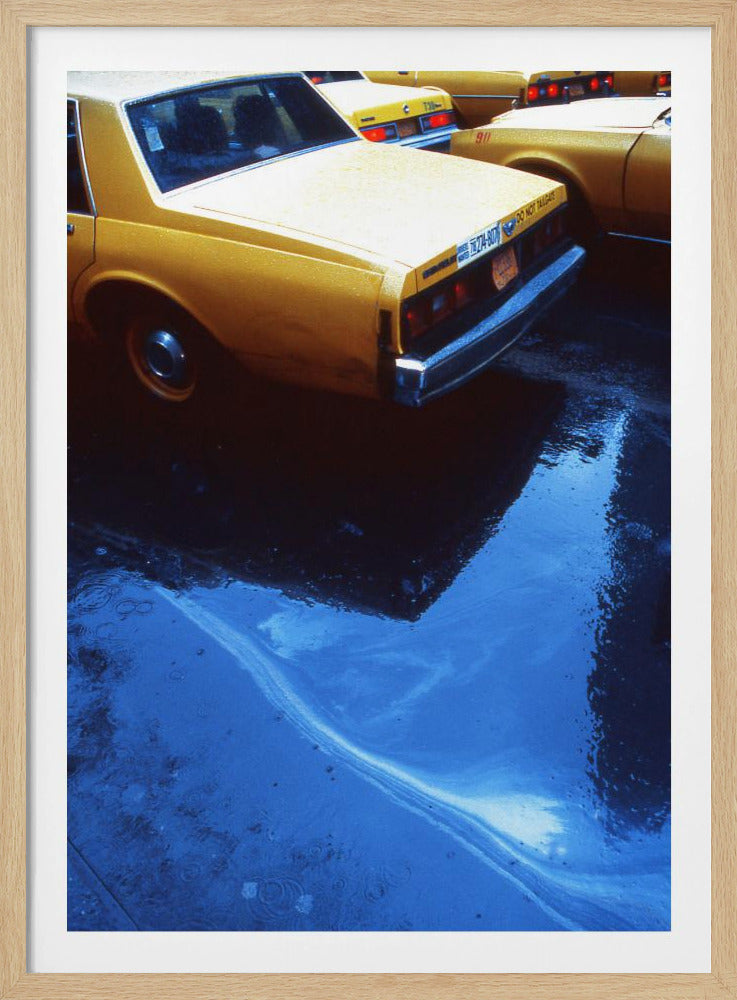 A close-up, angled view of the rear of a yellow taxi cab on a wet street. The car's dark reflection is cast upon a large, vibrant blue puddle in the foreground, with visible ripples from the rain. Wall Art