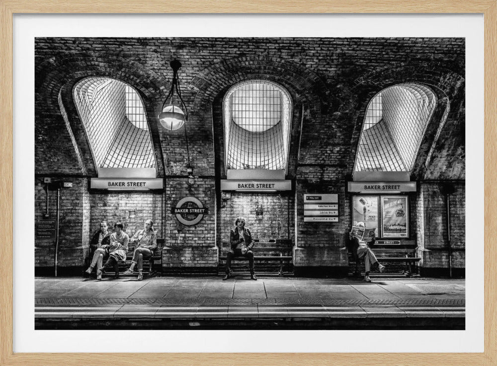 A framed, dramatic black and white photograph of the Baker Street underground station platform in London. Passengers wait on benches beneath iconic brick arches and large grated windows, with signs for 'Baker Street' and the London Underground roundel clearly visible. Print